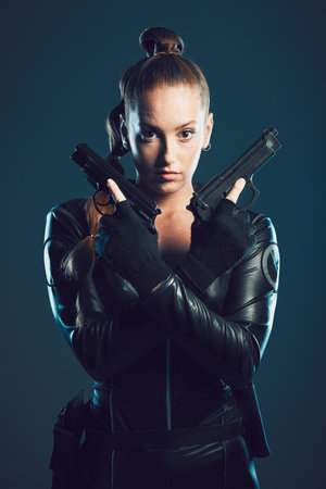 Woman, gun and assassin in studio portrait for costume, action or cyberpunk clothes. Girl, pistol and leather fashion for crime, gangster and futuristic spy with danger or future aesthetic with ninjaの写真素材