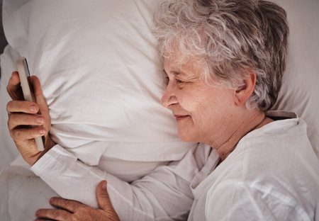 Smile, phone and senior woman in bed, text and read social media, message and relax in her home. Happy, elderly lady and online communication after wake up, browse and email on smartphone in bedroomの写真素材