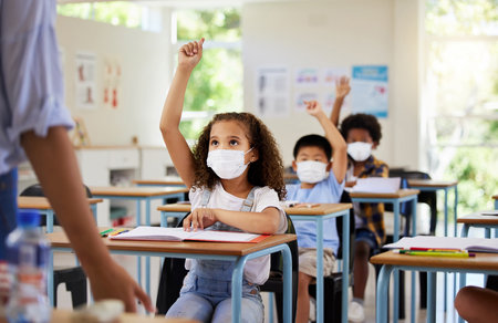 Covid, education and learning of eager, smart and clever children raising hands and wearing masks in a classroom. Teacher or educator asking questions for knowledge test at kids school in a pandemic.の写真素材