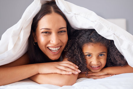 Children, family and bedroom with a girl and mother in bed under a blanket together for love or care in the home. Kids, trust and happy with a woman her daughter in the house to relax in the morningの写真素材