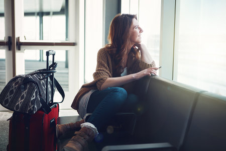 Time is getting close. a young woman sitting inside of an airport with her luggage and holding her cellphone while looking outside.の写真素材