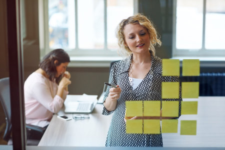 Making plans for success. a businesswoman reading adhesive notes on a glass wall with a colleague in the background.の写真素材