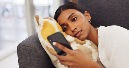 Social media phone, relax woman and reading chat on mobile app on the sofa in home, communication on internet and connection online with smartphone on couch. Young Indian girl on web with techの写真素材
