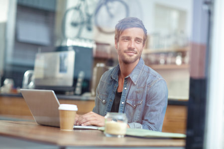 Relaxing with a fresh cup of coffee. a young man looking thoughtful while sitting with his laptop in a coffee shop.の写真素材