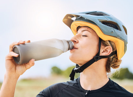 Woman drinking water, bottle and cycling with exercise outdoor, helmet for safety and health with hydration. Athlete, biking and female with healthy and active lifestyle, fitness and sports trainingの写真素材
