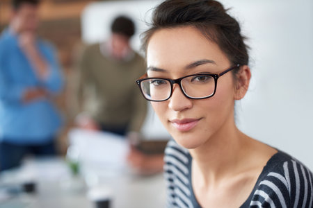 Starting my career from the ground up. Portrait of an attractive young woman sitting in an office with colleagues in the background.の写真素材
