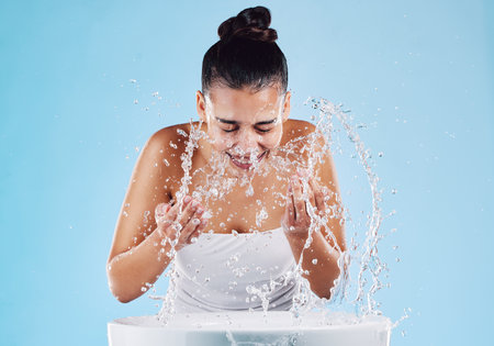 Sink, water splash and skincare of a woman washing face for natural beauty and facial wellness. Isolated, blue background and studio with a female model doing dermatology and cosmetic routineの写真素材