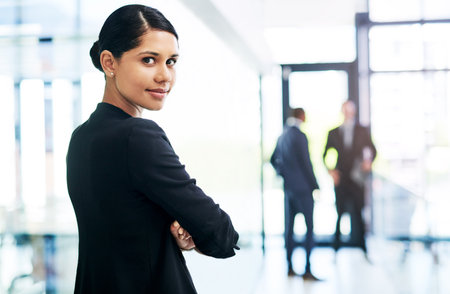 Watch me take over this company. Cropped portrait of an attractive young businesswoman standing with her arms folded in an office with her colleagues in the background.の写真素材