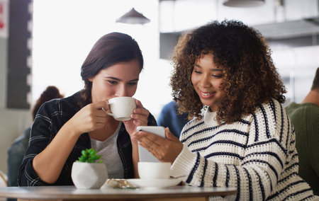 Look at the cute message he just sent me. two young women looking at something on a cellphone in a cafe.の写真素材