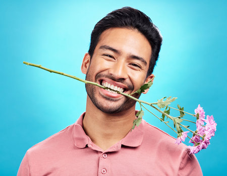 Teeth, flower and happy with portrait of man in studio for celebration, gift and romance. Funny, goofy and present with male isolated on blue background for happiness, smile and valentines day mockupの写真素材
