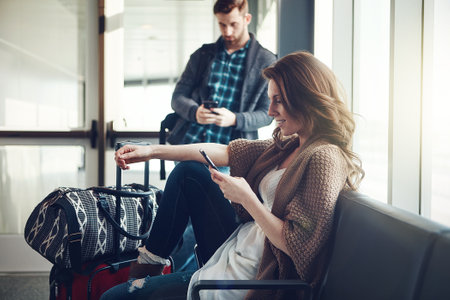 Just saying goodbye to the family. a young couple sitting in an airport with their luggage while making use of their cellphones.の写真素材