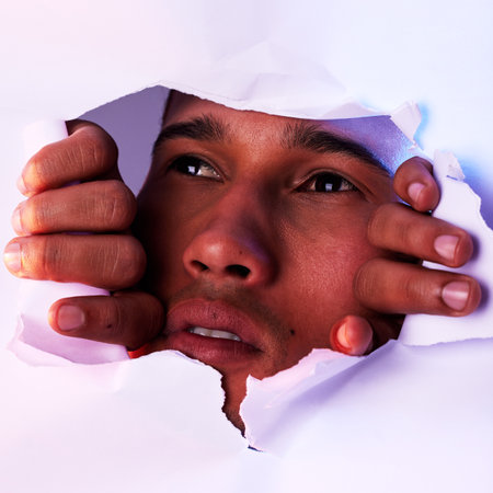 Face, wall and a man looking through a hole to discover, explore or search while on an adventure. Head, eyes and thinking with a handsome young male closeup in studio to look out from a tear or gapの写真素材