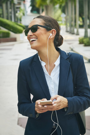 Looking forward to our meeting. a young businesswoman her phone and earphones while out in the city.の写真素材