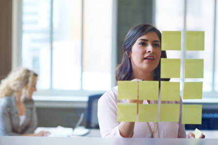 Streamlining ideas. a businesswoman reading adhesive notes on a glass wall with a colleague in the background.の写真素材