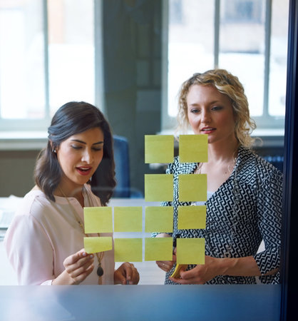 This one looks good. two businesswomen brainstorming with adhesive notes on a glass wall in an office.の写真素材