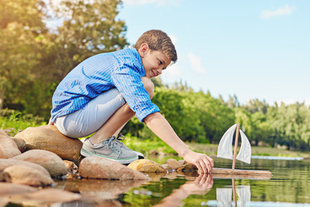 Its her maiden voyage. a young boy playing with a toy boat by the water.の写真素材