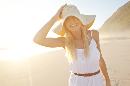 Beauty in the sunlight. a gorgeous young woman wearing a white dress and sunhat on the beach.の写真素材