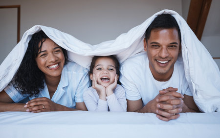 Happy, love and portrait of a family on a bed in their bedroom relaxing and bonding together. Happiness, smile and girl child laying with her mother and father with a blanket in their modern home.の写真素材