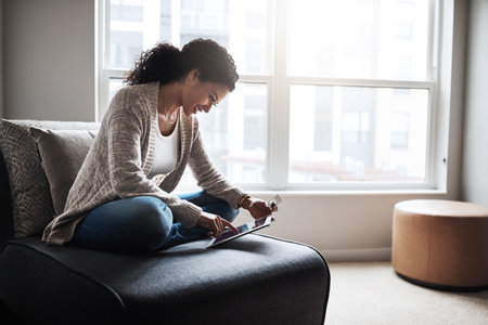 This is too easy. a cheerful young woman relaxing on a chair while doing online shopping on a digital tablet at home.の写真素材