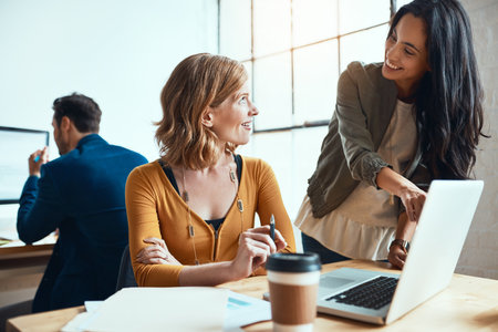 Tell me more about this. two young businesswomen working together in their modern office with a male colleague in the background.の写真素材