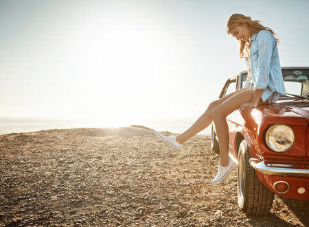 Summertime is road trip time. a beautiful young woman relaxing on the hood of her car on a road trip.の写真素材