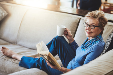The retired life aka the good life. Portrait of a senior woman relaxing with a warm beverage and a book on the sofa at home.の写真素材