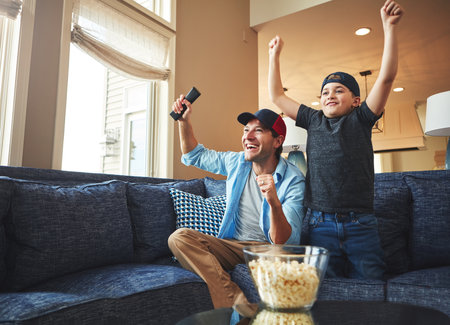 Building that father son bond through sports. an enthusiastic father and son watching a sports match on tv at home.の写真素材