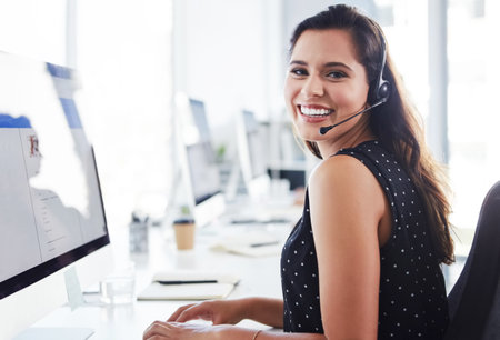 Ready and available to take your call. a young woman using a computer and headset in a modern office.の写真素材