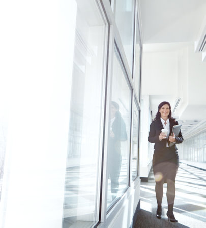 Busy businesswoman walking to her office holding files and drinking coffee. One female lawyer carrying documents and a disposable cup at work in the hallway. Executive professional at a startupの写真素材