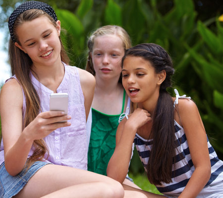 Catching up on the latest gossip online. A young girl showing her friends something on a cellphone while theyre sitting outside.の写真素材