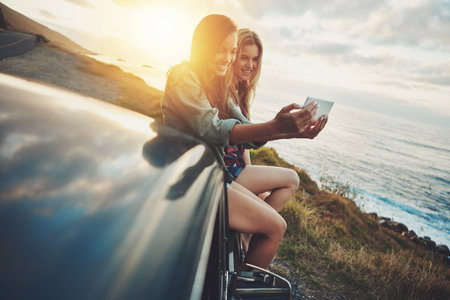 One more for the road. Full length shot of two friends taking a selfie while sitting on the hood of a car.の写真素材