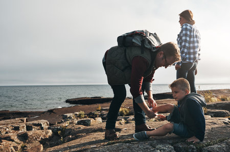 Lets take a little break here. a cheerful man helping to put his sons shoes on while standing next to the ocean outside during the day.の写真素材