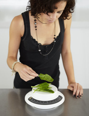 Woman, scale and spinach in kitchen for food, diet or eating disorder with leaves for meal plan. Girl, mental health and vegetable leaf to lose weight with anorexia, bulimia or starving in houseの写真素材