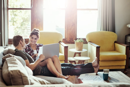 Getting things done in comfort. a young couple using a laptop and going through paperwork together on the sofa at home.の写真素材