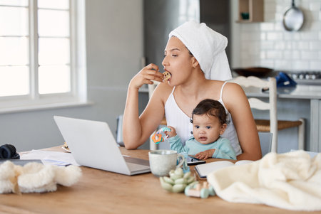 Hope youre having a good morning because I have a busy one. Shot of a woman eating rusks while working on her laptop and holding her baby on her lap.の写真素材