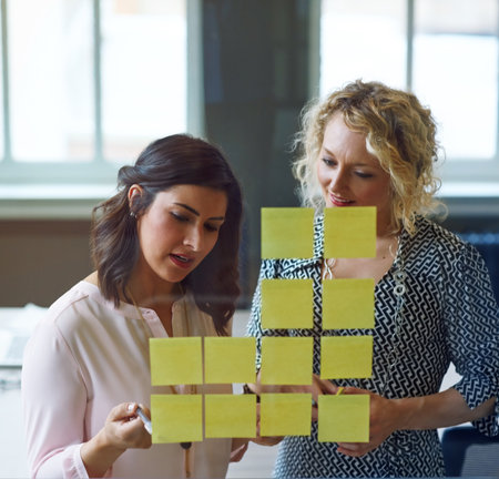 Theyre all about great ideas. two businesswomen brainstorming with adhesive notes on a glass wall in an office.の写真素材