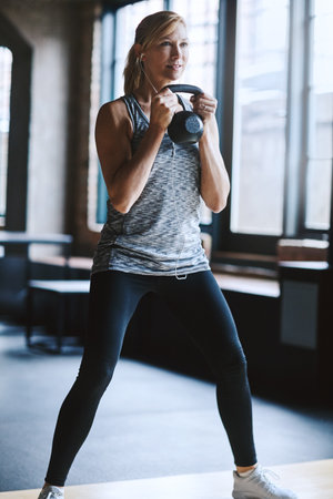 Raise your game. an attractive young woman listening to music while working out with a kettle bell in the gym.の写真素材