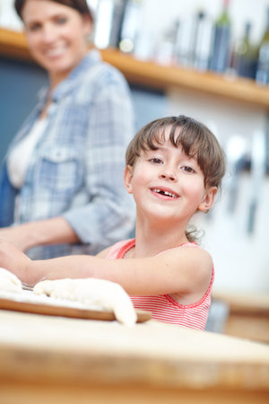 What can I bake for you today. A cute little girl working with cookie dough with her mother standing in the background.の写真素材