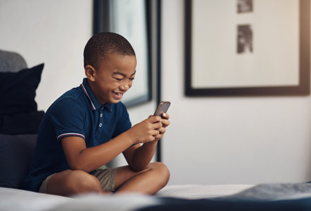 Mom gave me her old cellphone for gaming. a young boy using a cellphone while sitting on his bed.の写真素材
