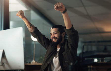 Putting in the hours gets you that perfect score. a young businessman cheering while using a computer during a late night at work.の写真素材