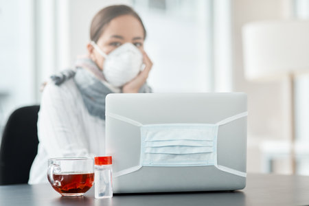 These are difficult times, but we can do this. a masked young businesswoman working with hand sanitiser, a laptop and herbal tea at her desk.の写真素材