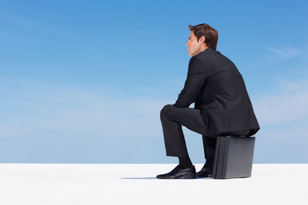 Thoughtful business man sitting on briefcase against blue sky. Thoughtful young business man sitting on briefcase against clear sky.の写真素材