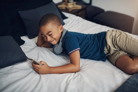 I get to enjoy screen time until 6pm. a young boy using a cellphone while lying on his bed.の写真素材