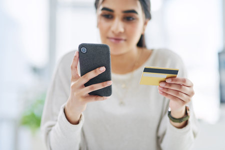 Doing some shopping from the business budget. a young businesswoman using a cellphone and credit card in an office.の写真素材