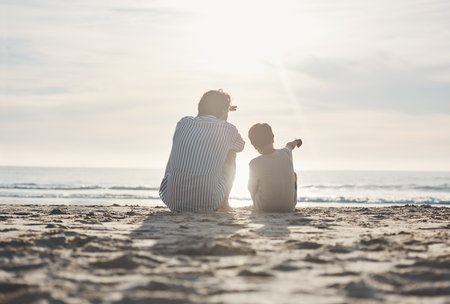 Dad, look over there. Rearview shot of an unrecognizable father sitting and bonding with his son during an enjoyable day on the beach.の写真素材