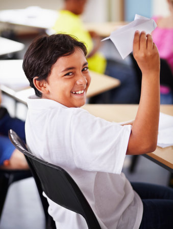 Portrait, student and paper plane in classroom and happy, learning at desks or Indian boy, play and origami jet or class distraction. Playful smile, kid and holding airplane and middle school funの写真素材