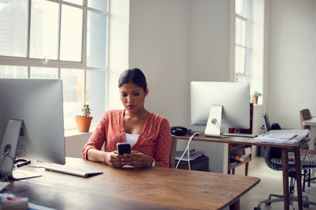 Keeping her clients close to her. a young businesswoman using a mobile phone at her desk in a modern office.の写真素材