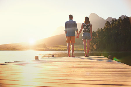 Love under a beautiful sunset. Rearview shot of an affectionate young couple standing on a dock at sunset.の写真素材