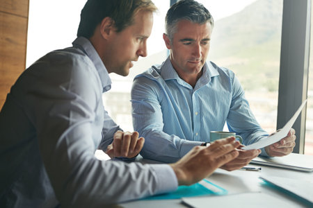 Putting ideas to paper. two businessmen discussing paperwork while sitting in an office.の写真素材