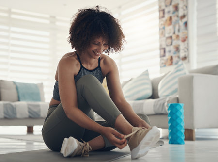 Lets get this quarantine body moving. an attractive young woman sitting alone in her living room and tying her shoelaces before working out.の写真素材